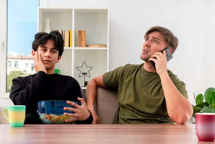 Unpleased Young Blonde Handsome Man Talking Phone Sitting Table With Disappointed Young Brunette Handsome Guy Putting Hand Face Holding Bowl Chips Inside Design Living Room 141793 70085