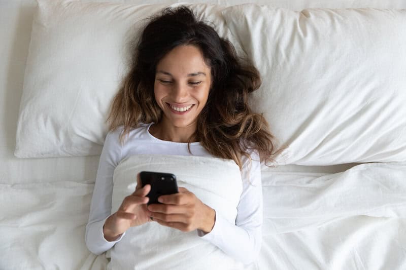 Shutterstock Woman Happily Texting On Phone In Bed Smiling 052220